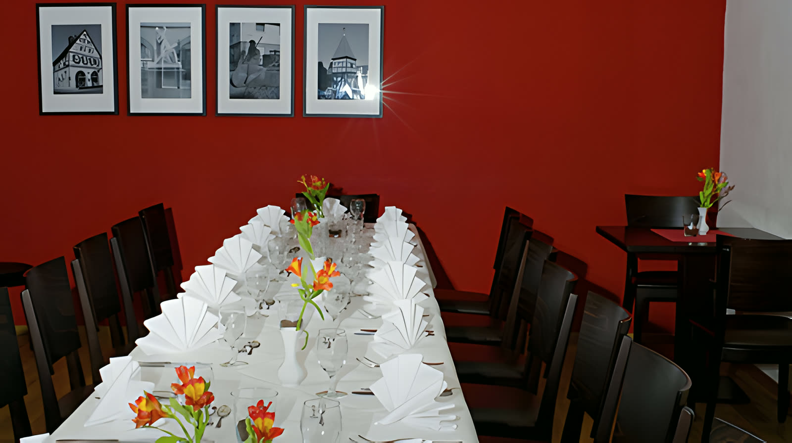 Nebenzimmer mit tiefroter Wand, langem festlich eingedecktem Tisch und orangenen Lilien